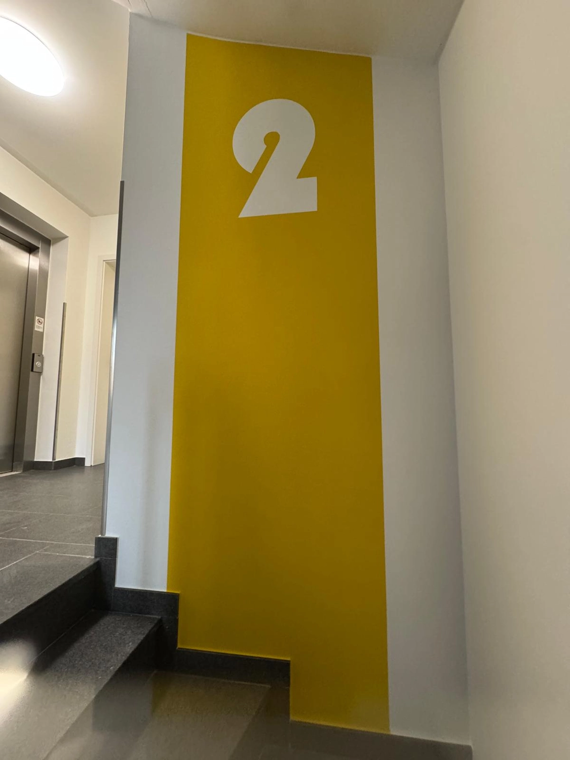 Karl Hansel Inh – Baudekoration – Eine leuchtend gelbe Wand mit einer großen weißen „2“ im Flur eines modernen Gebäudes neben einer Treppe und einem Aufzug.
