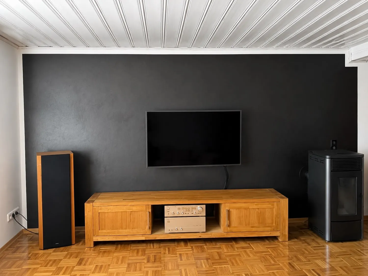 Karl Hansel Inh – Baudekoration – Ein minimalistisch gestaltetes Wohnzimmer mit einem an der Wand montierten Fernseher über einer Medienkonsole aus hellem Holz, flankiert von einem Standlautsprecher und einem Pelletofen vor einer dunklen Akzentwand.