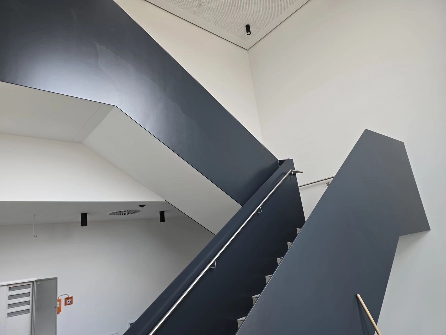 Karl Hansel Inh – Baudekoration – Eine moderne, minimalistische Treppe mit dunkelgrauen Geländern und weißen Wänden unter heller Innenbeleuchtung.