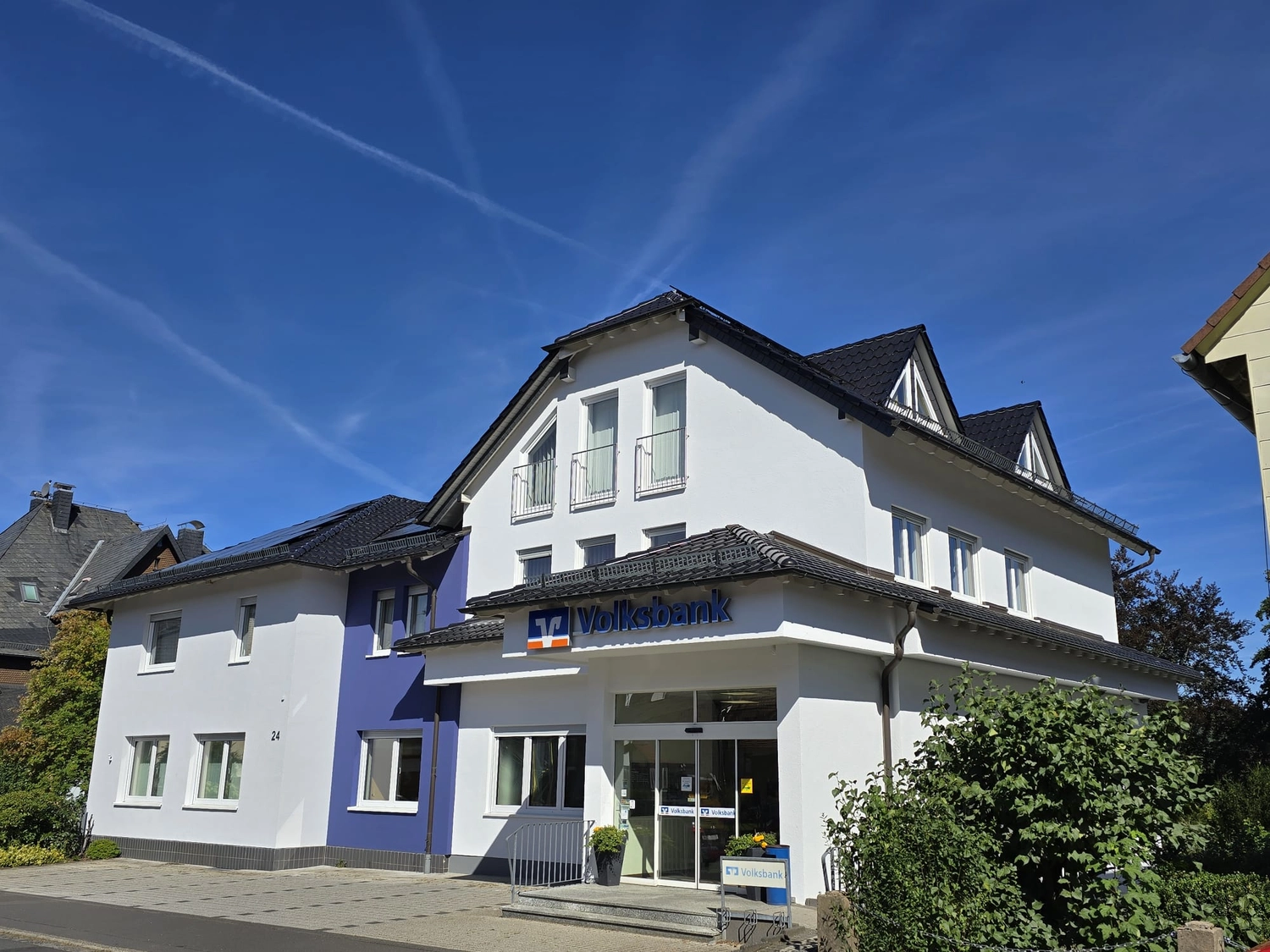 Karl Hansel Inh – Baudekoration – Ein strahlend weißes Volksbank-Gebäude mit einem dunklen Ziegeldach vor strahlend blauem Himmel.