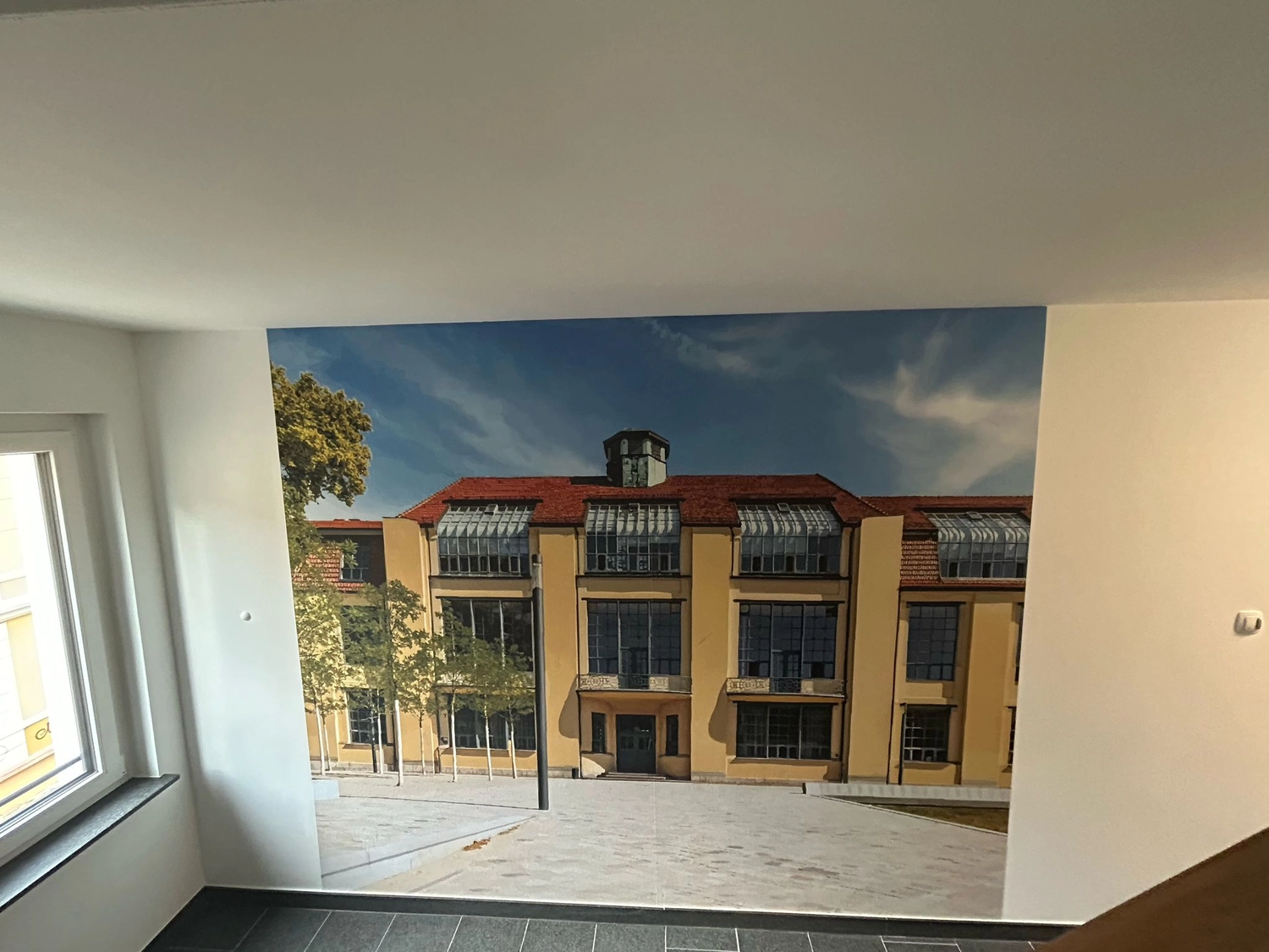 Karl Hansel Inh – Baudekoration – Eine große Wandtapete in einem Raum, die ein gelbes Gebäude mit rotem Dach unter blauem Himmel zeigt.